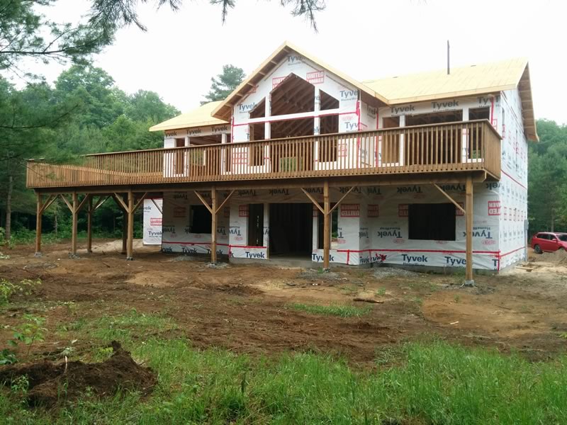 A raised bungalow on a cleared lot with wood-framed walls in white house wrap and a long deck on posts