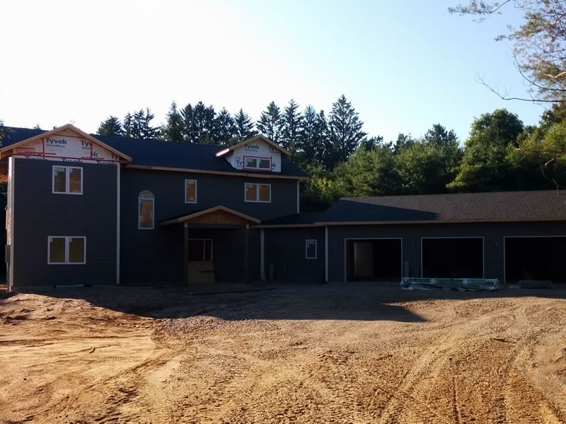 A nearly finished two-storey house with dark siding and a long attached garage, behind a wide yard of bare, freshly graded sandy soil marked with tire tracks