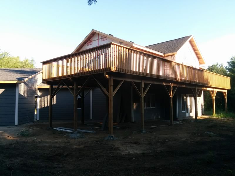 The back of a raised bungalow under construction, a long wood deck on posts above a walkout lower level with patio doors at ground level