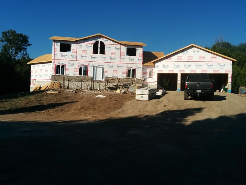 A two-storey house in white house wrap with an attached three-bay garage under construction, a pickup truck in the open bay and fresh fill in the yard