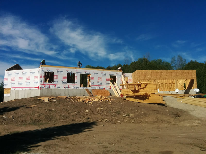 A rural build site with bare excavated soil in the foreground, a new house under construction with its walls framed and wrapped, and a line of evergreens behind