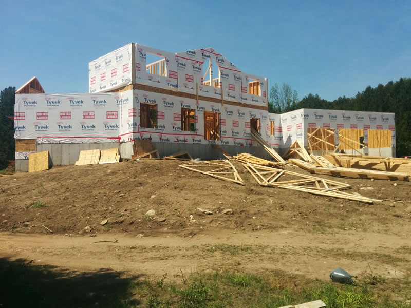 A large house under construction, wrapped in house wrap, standing behind a stretch of excavated earth with offcuts of lumber and stacked materials in the foreground