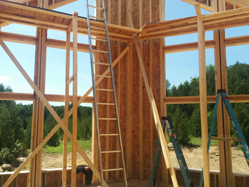 Open wood-framed exterior wall with studs, sheathing and a ladder, trees visible through the window openings