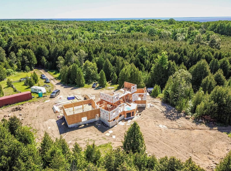 Aerial view of a rural build site: a framed house in a cleared sandy area surrounded by dense evergreen forest, with a long gravel access lane, parked vehicles and a storage container to one side