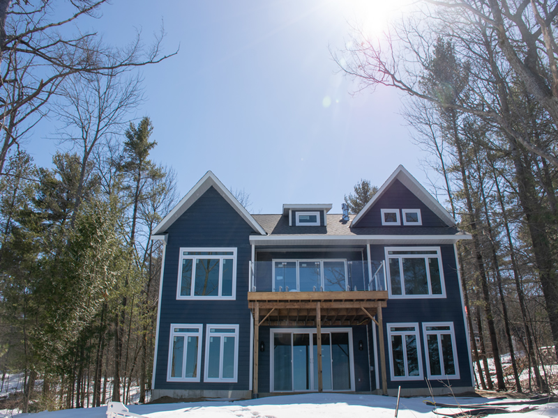 The Crandon in Muskoka — a navy-sided custom cottage with twin gables and a glass-railed balcony over a timber-post walkout, on a snowy treed lot