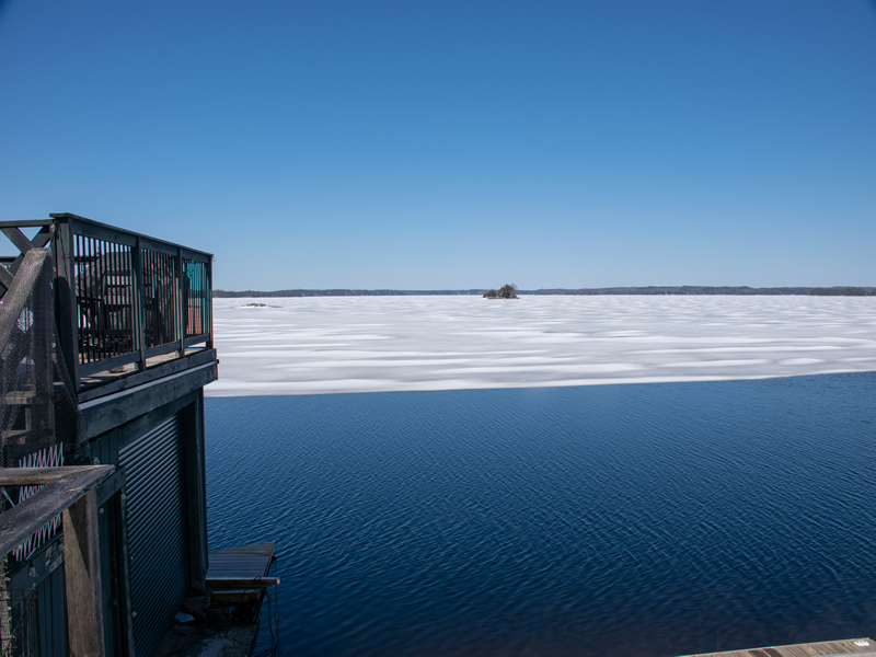 View past the corner of a boathouse deck to a lake where open blue water meets a wide sheet of white ice that still covers most of the bay