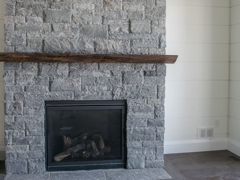A floor-to-ceiling grey stone fireplace with a rough timber mantel and a glass-fronted firebox, in a room with white shiplap walls