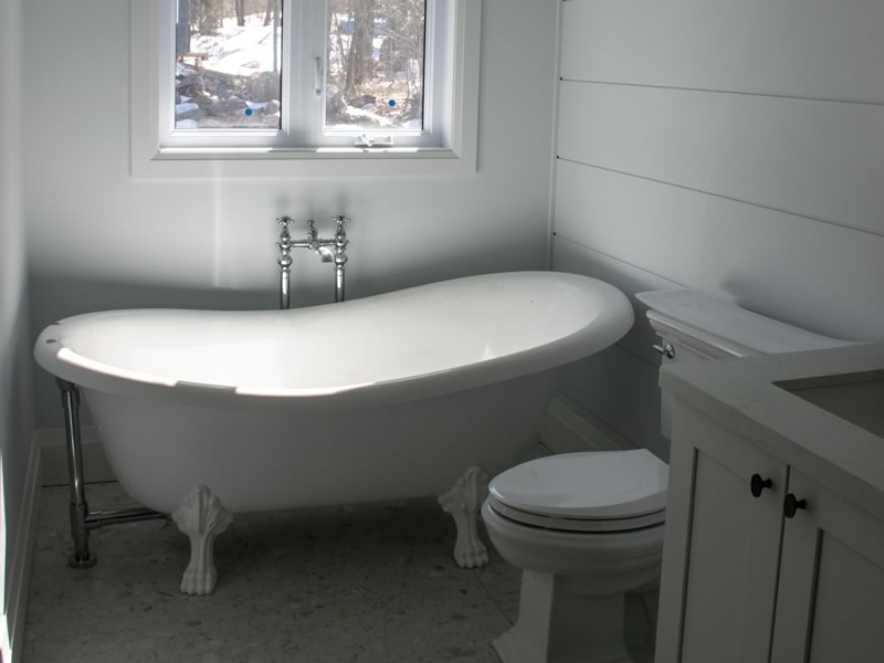 A cottage bathroom with white shiplap walls, a clawfoot tub under a window with snow-covered ground outside, and a toilet beside a white vanity