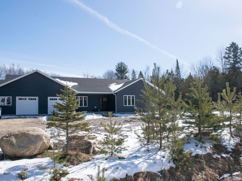 A grey single-storey home with a two-car garage seen in winter across a snowy, rocky slope, with a large boulder and young pines in the foreground