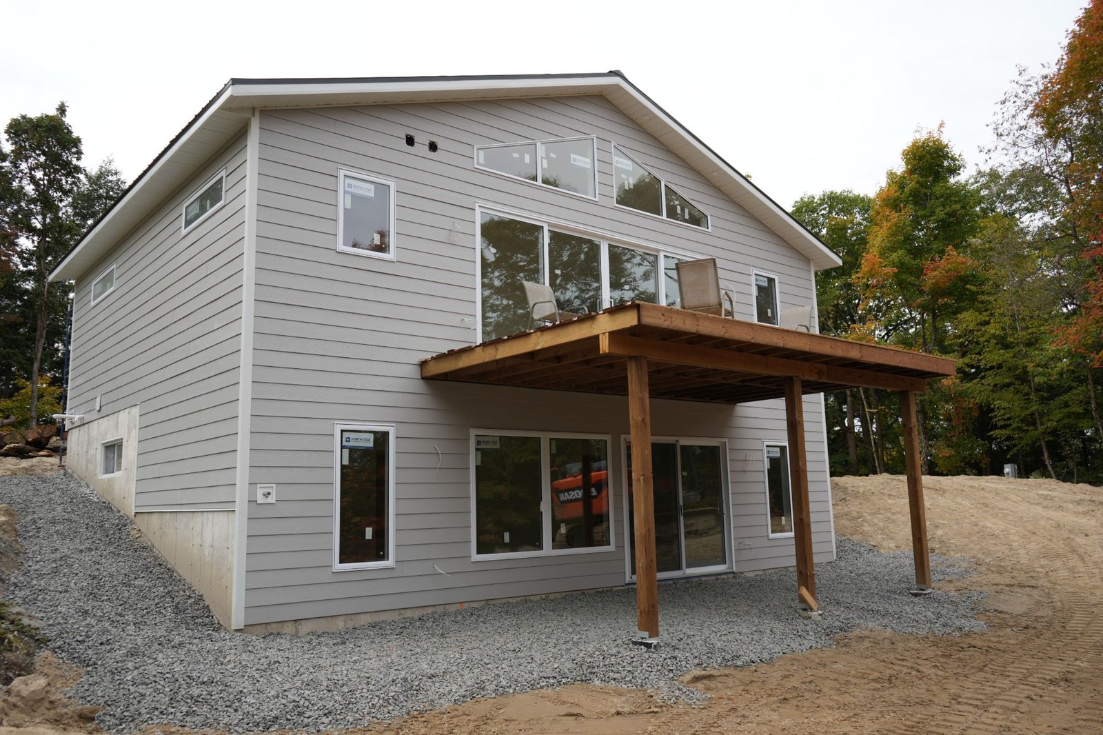 Rear of a grey-sided house under construction on a sloped lot, the concrete foundation wall stepping down with the ground to a walkout level, deck posts on their own footings and crushed stone laid against the wall