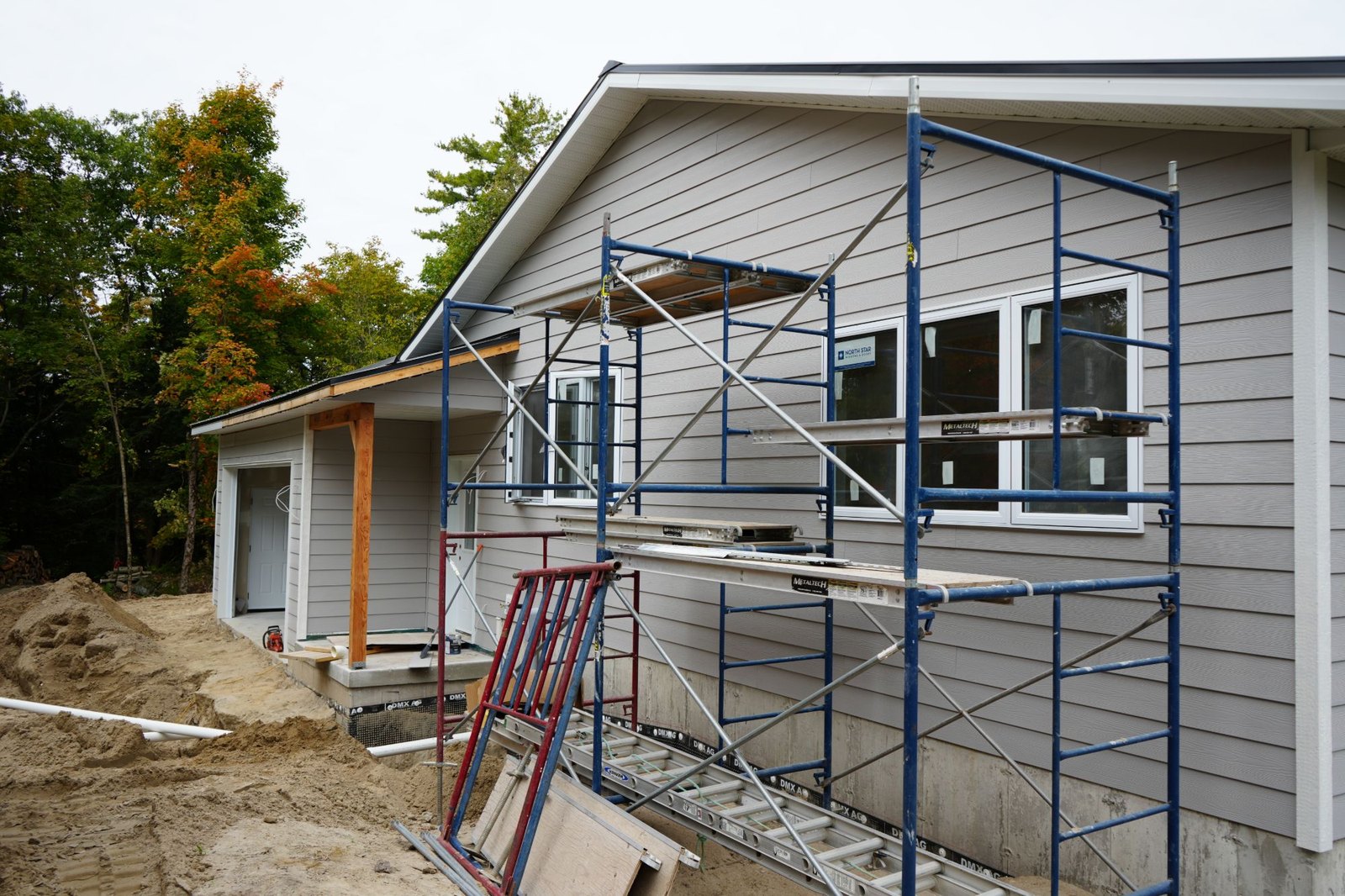 Side of a grey-sided house under construction with scaffolding against the wall, the concrete foundation below wrapped in dimpled membrane and an open trench of sand with drain pipe running beside it