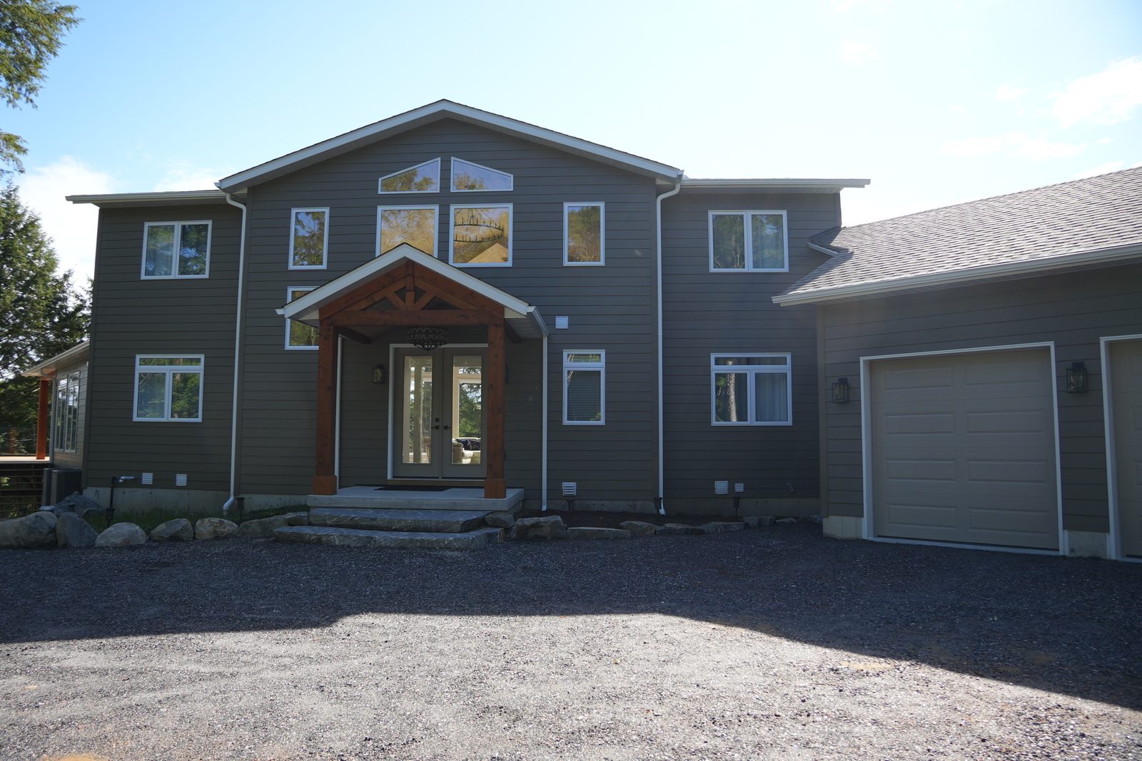 Front of a finished two-storey house with taupe siding, a timber-frame entry porch and an attached garage, set on a wide gravel driveway with trees behind