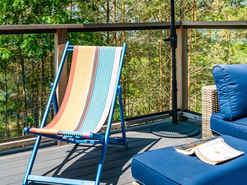 A striped sling chair and blue outdoor cushions on a grey composite deck, behind a railing of clear glass panels set between wood posts with trees beyond