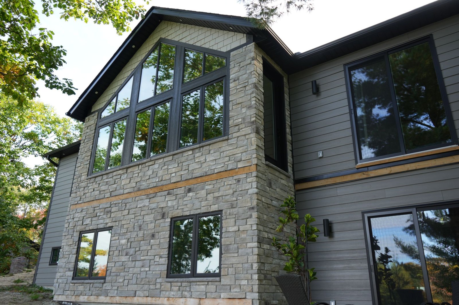 Corner of a home with a stone-faced gable wall, a tall window wall and grey siding, stone being weight that the wall below carries down to its footing