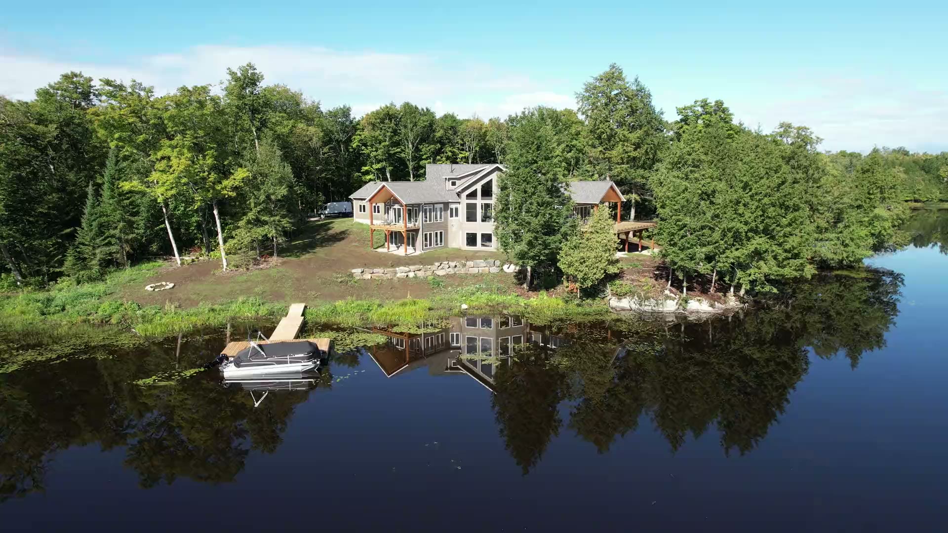Aerial view of a finished lakeside home set back from the shore on a wooded lot, with a cleared lawn, an armour-stone wall, a short dock and a pontoon boat on still water